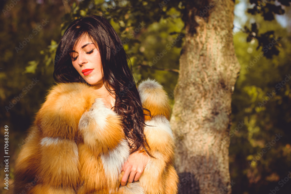 Luxurious brunette young woman with curly hair in a red fox fur coat in the autumn forest. The beauty of natural fur. Luxurious brunette young woman with curly hair in a red fox fur coat in the autumn forest. The beauty of natural fur.