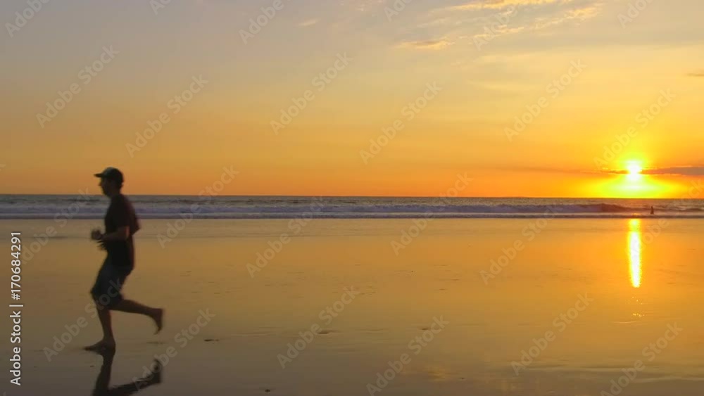 SLOW MOTION, CLOSE UP: Young man running barefoot along smooth sandy beach. Ocean waves splashing waterdrops at dramatic golden light sunset. Guy in summer clothes jogging along the seashore at dusk SLOW MOTION, CLOSE UP: Young man running barefoot along smooth sandy beach. Ocean waves splashing waterdrops at dramatic golden light sunset. Guy in summer clothes jogging along the seashore at dusk