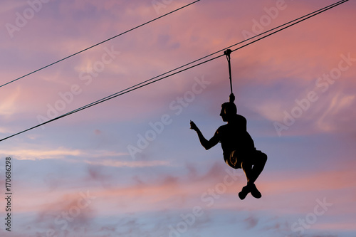 A man is jumping on a rope swing and removing video on the smartphone at the same time. Silhouette on the background of the evening sky.
