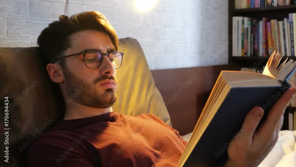 Young handsome man lying in bed and reading a book in his bedroom at home at night