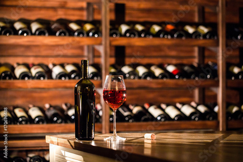 Bottle and glass filled with red wine placed on a wooden table. Wine vault location.