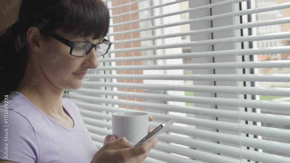 Young woman in glasses drinks coffee standing with smartphone by window in the office. Girl looks out the window and uses a smartphone.Text message, online surfing, app, social network. Young woman in glasses drinks coffee standing with smartphone by window in the office. Girl looks out the window and uses a smartphone.Text message, online surfing, app, social network.
