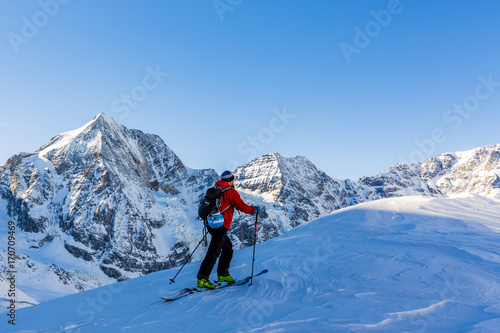Mountaineer backcountry ski walking up along a snowy ridge with skis in the backpack. In background blue sky and shiny sun and Zebru, Ortler in South Tirol, Italy. Adventure winter extreme sport.