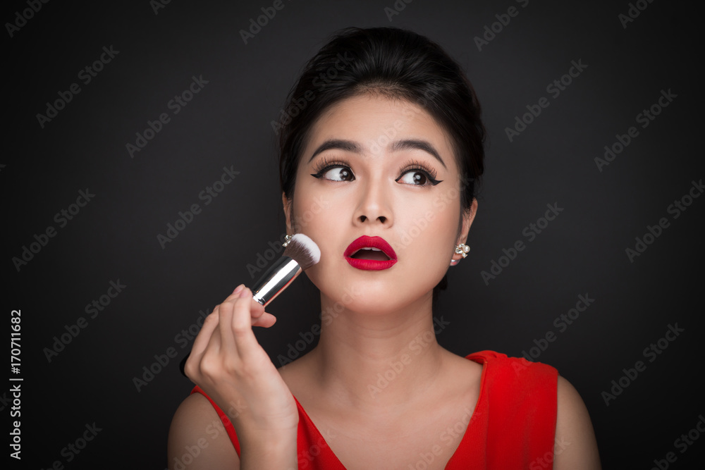 Fototapeta premium Cosmetic powder brush. Asian woman applying blusher on her cheeks with perfect make-up and red lips