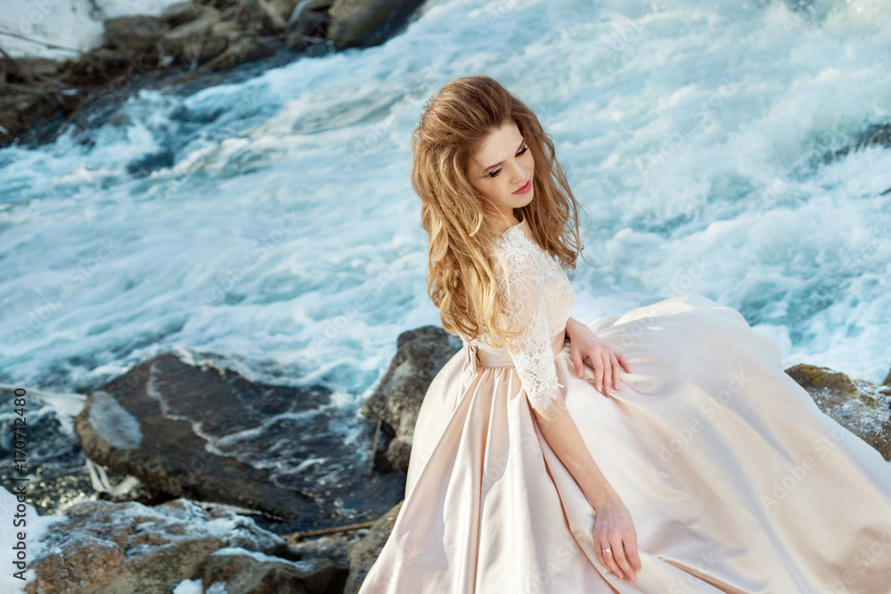 Beautiful young girl in lush bride's dress with hair and makeup on a background of spring nature near small river and forest