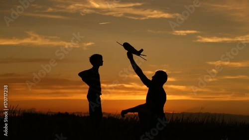 A happy family. The boy on his father's shoulders is playing with an airplane toy. Silhouette at sunset by the lake. at this time a real airplane is flying in the sky