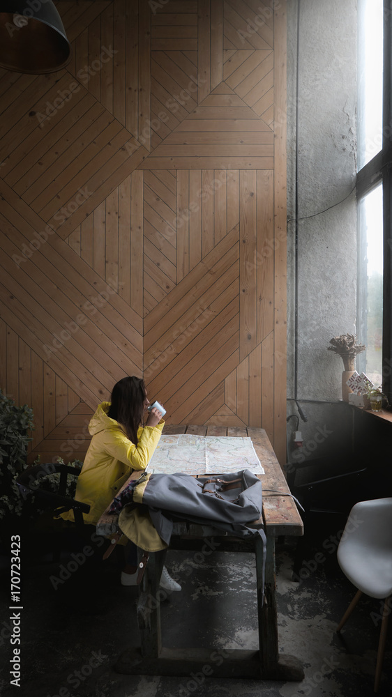 Fototapeta premium Beautiful Caucasian female millennial hiker tourist in a yellow raincoat studying a map in a rustic lodge