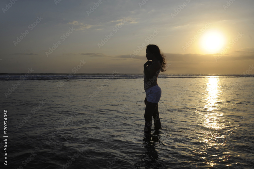 Silhouette of young beautiful asian woman standing on water free and relaxed looking at horizon on sunset beach in Bali