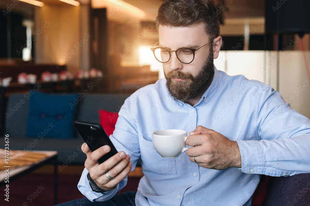 Young serious attractive bearded businessman in blue shirt and glasses sits on sofa in room, drinks coffee and uses smartphone Young serious attractive bearded businessman in blue shirt and glasses sits on sofa in room, drinks coffee and uses smartphone