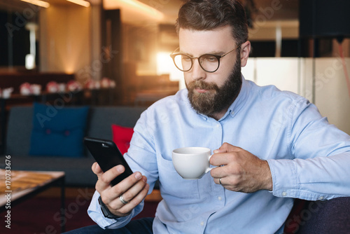Young serious attractive bearded businessman in blue shirt and glasses sits on sofa in room  drinks coffee and uses smartphone
