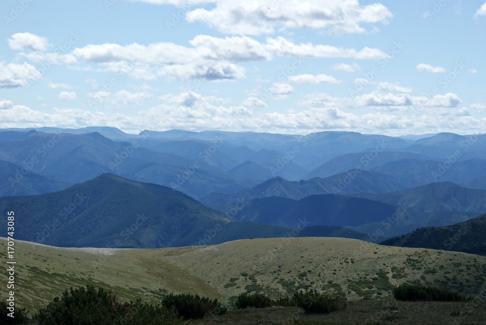 Fototapeta premium A mountain landscape shot from one of the peaks. Blue mountains to the horizon and the sky with clouds. Chamar-Daban (Khamar-Daban) montains, Siberia, Russia. Summer, day.