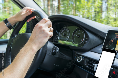 Male hands holding car steering wheel. Hands on steering wheel of a car driving near the lake. Man driving a car inside cabin. Smartphone in holder with isolated white empty blank screen. Copy space