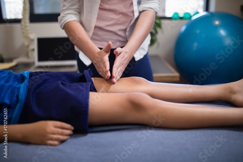 Young female therapist massaging knee of boy lying on bed