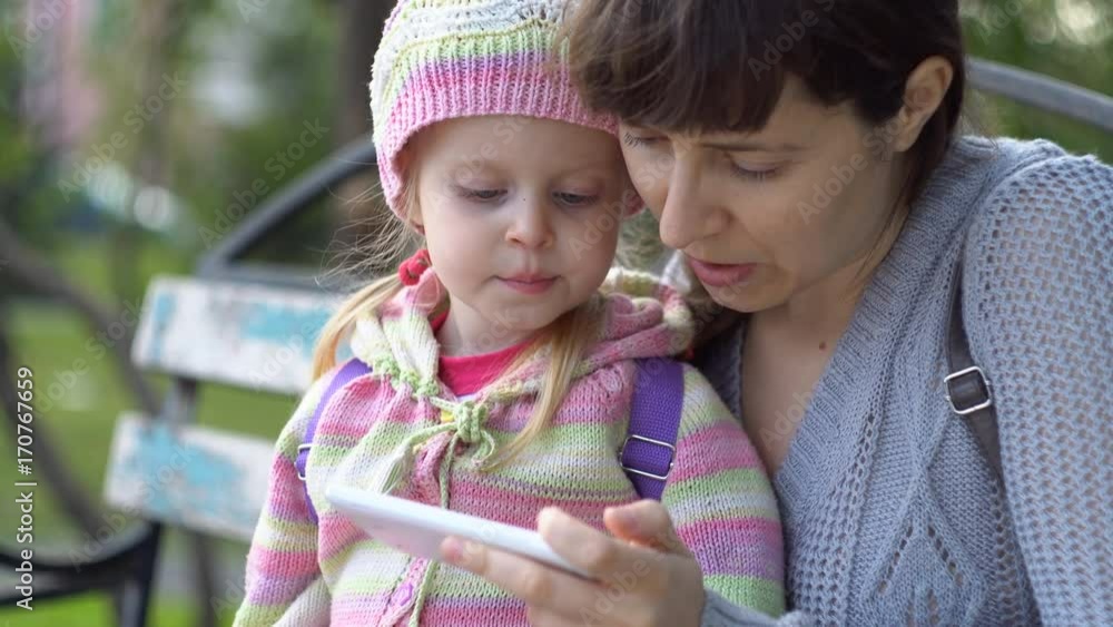 Mother with her little daughter dressed in a striped sweater together use the phone sitting on a bench in a public park on a cool evening, close-up shot. Mother with her little daughter dressed in a striped sweater together use the phone sitting on a bench in a public park on a cool evening, close-up shot.