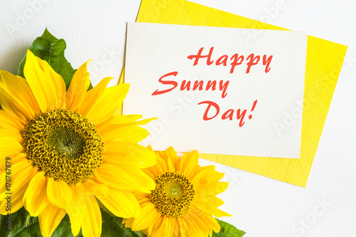 Greeting card with text happy sunny day and with big beautiful yellow sunflowers on the white table. Positive emotions.