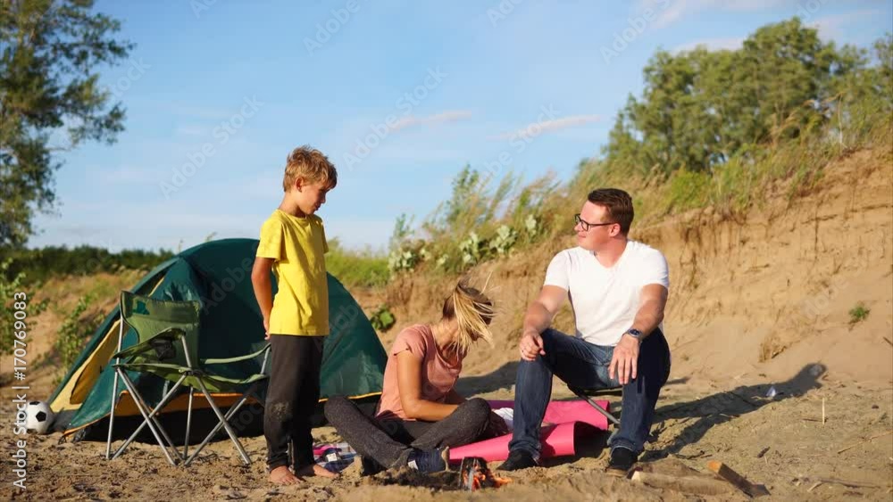 A young and friendly family who spend weekends in nature, sit near a campfire and a tent with their son, mother sits on a sandy beach, a boy stands and looks at the fire the husband talks to the child A young and friendly family who spend weekends in nature, sit near a campfire and a tent with their son, mother sits on a sandy beach, a boy stands and looks at the fire the husband talks to the child