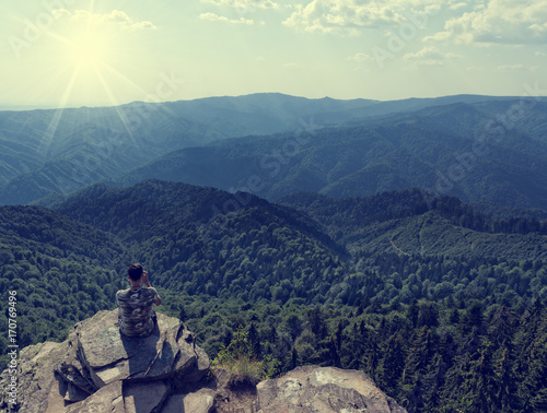 Young person standing on a rock above mountain peaks covered with pine forest and a beautiful summer day with a blue sky and the shining sun. Concept for meditation or loneliness 