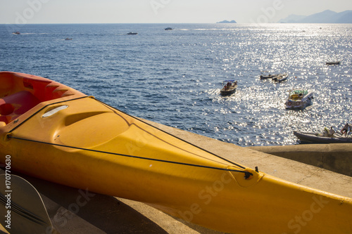 Dettaglio di una canoa gialla monoposto ferma sulla spiaggia. Nel mare gommoni, barche e moto d'acqua sfrecciano verso l'orizzonte dove si intravede l'isola di Capri.