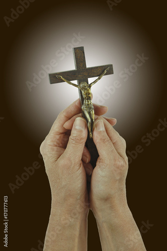 Praise the Lord - Female hands holding a small statue of Christ on the Cross, against a dark brown background with white light behind the cross 