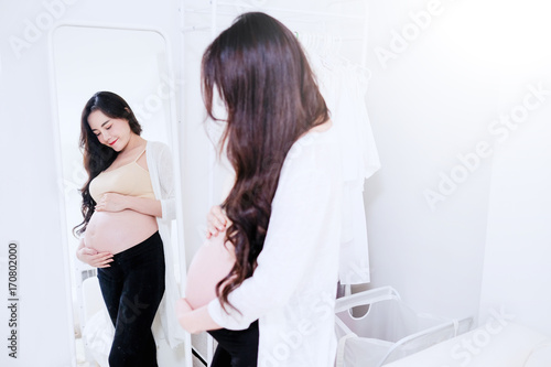 Young pregnant woman standing in front of the  in a mirror while touching bell ; background of the white bedroom photo