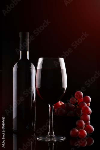 Bottle of red wine, a glass of red wine and grapes on a dark background.