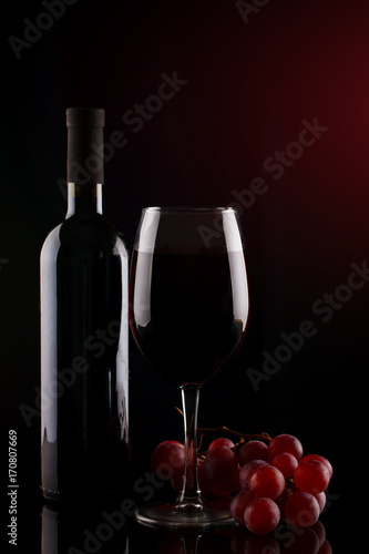 Bottle of red wine, a glass of red wine and grapes on a dark background.