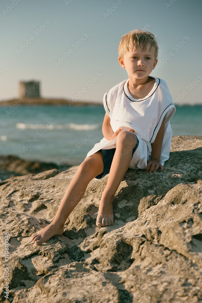 boy dressed in a large shirt seat on a stone,  La Pelosa Beach on the island Sardinia.