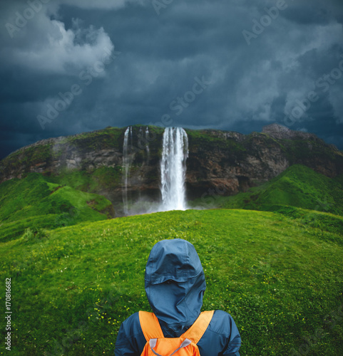 Girl in waterproof clothing stands under the Seljalandsfoss waterfall in Iceland. back view, woman with small orange backpack