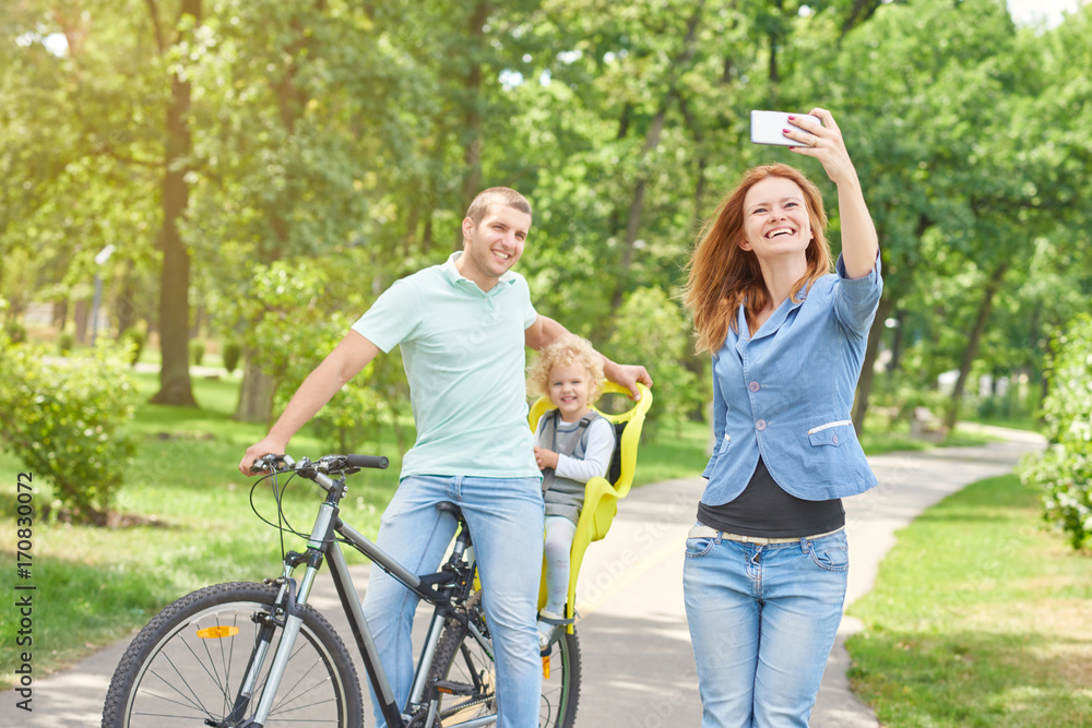 Beautiful young woman taking a selfie with her husband and baby on bicycle while relaxing outdoors at the park on a sunny warm day memories emotions parenting parents recreation activity sports. Beautiful young woman taking a selfie with her husband and baby on bicycle while relaxing outdoors at the park on a sunny warm day memories emotions parenting parents recreation activity sports.