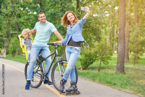 Full length shot of a young woman wearing rollerblades posing with her husband and baby on bicycle taking a selfie using smart phone at the park copyspace. © serhiibobyk