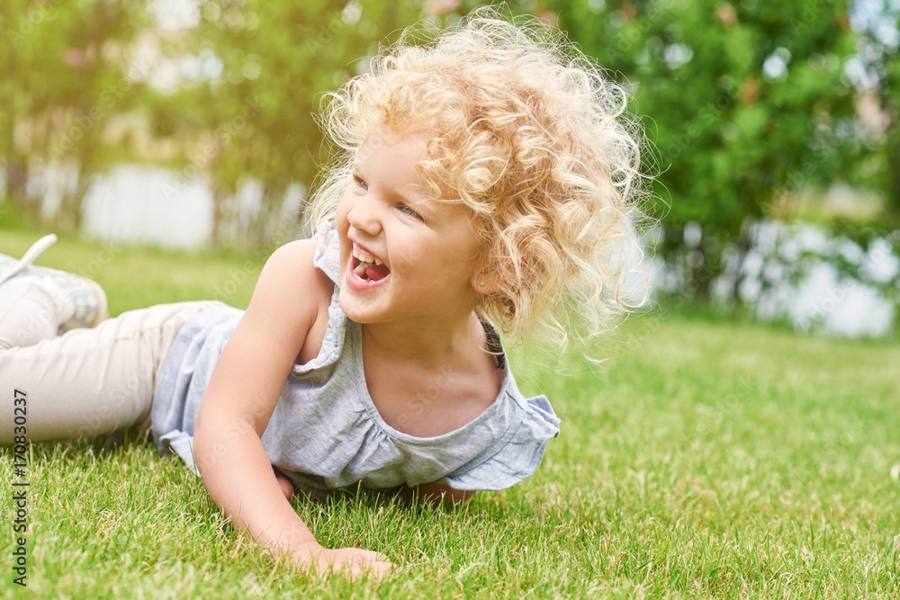 Fototapeta premium Shot of a cute little girl with blonde curly angelic hair lying on the grass laughing happily looking away copyspace nature enjoyment emotions kids children family recreation summer concept.
