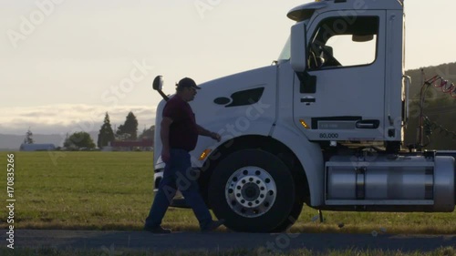 Driver walks up to truck parked on rural road, climbs up to cab and shuts door in late afternoon. Side view, slow motion 4K recorded at 60fps