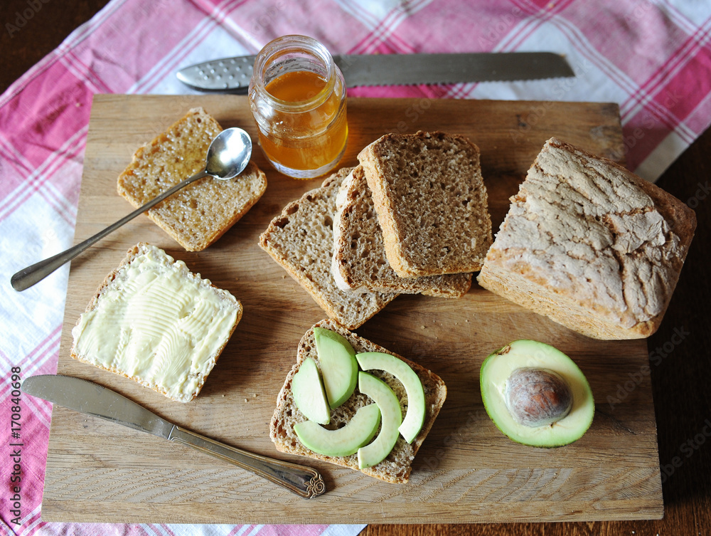 Healthy breakfast with fresh bread, avocado, honey jam and butter on a wooden board on plaid tablecloth