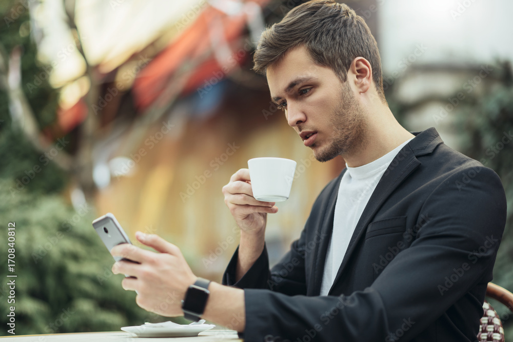 Modern technology, business, communication and people concept. Confident stylish Caucasian businessman in suit drinking coffee at terrace, reading news from his device smart phone outdoor