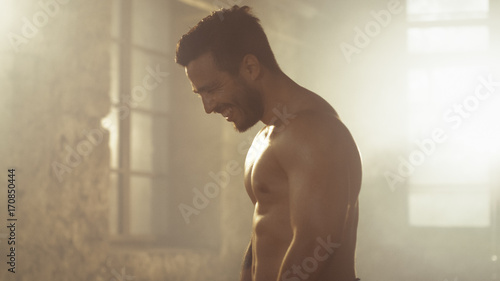 Handsome Shirtless Man with Naked Muscular Torso with Visible Six Pack Stands Resting after Bodybuilding Exercise. He's in the Middle of Abandoned Factory Building.