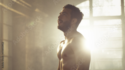 Handsome Shirtless Man with Naked Muscular Torso with Visible Six Pack Stands Resting after Bodybuilding Exercise, He Wipes Sweat from His Forehead. He's in the Middle of Abandoned Factory Building.