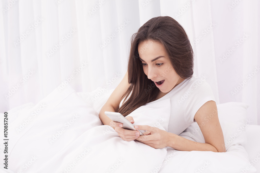Young happy beautiful brunette woman using the mobile phone in white bed. Young happy beautiful brunette woman using the mobile phone in white bed.
