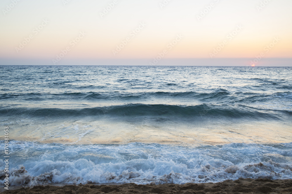 Vista dell'alba dalla spiaggia. Ilmare è calmo e le onde sono poche che si infrangono sulla battigia. Il sole all'orizzonte dona le tinte calde al paesaggio.