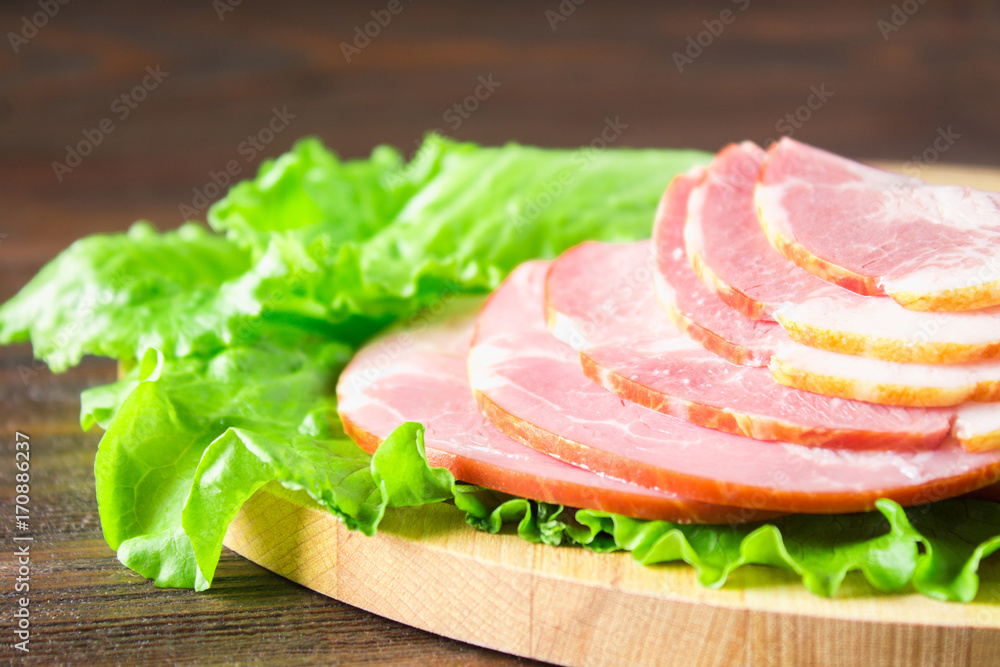 Sliced ham with fresh green lettuce leaves on a round cutting board. Meat products on a brown wooden table. Sliced ham with fresh green lettuce leaves on a round cutting board. Meat products on a brown wooden table.