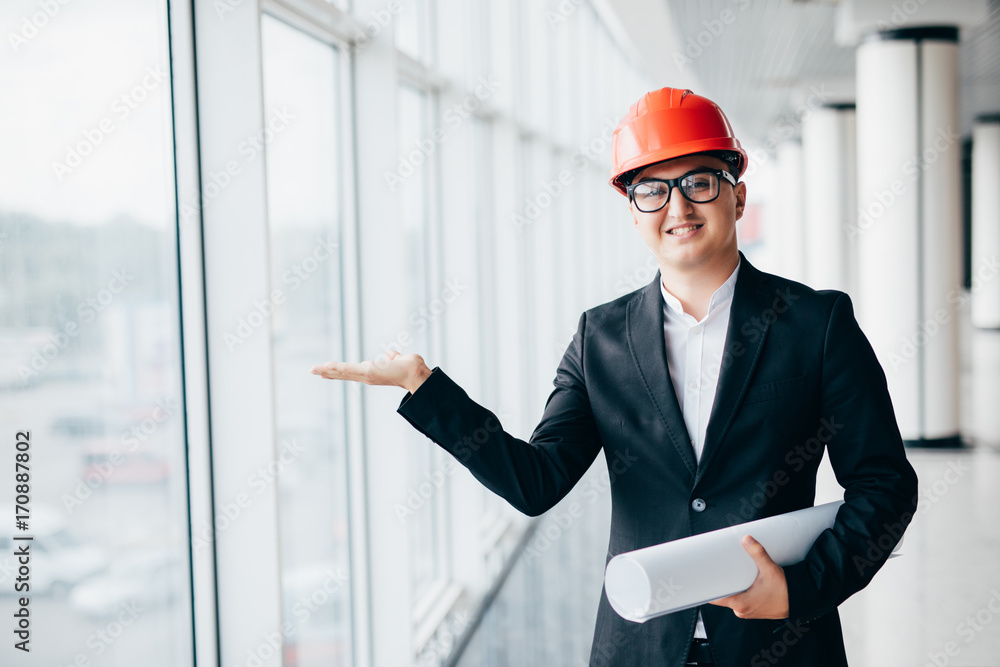 Man architect in protective hamlet looking at blueprints in hamlet with open hand for copyspace near panoramic windows Man architect in protective hamlet looking at blueprints in hamlet with open hand for copyspace near panoramic windows