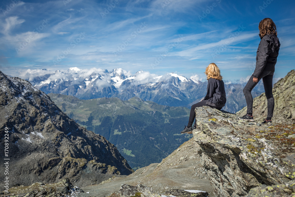 deux jeunes filles , une blonde et une brune , assise et debout, face à un paysage de montagne avec des cimes enneigées