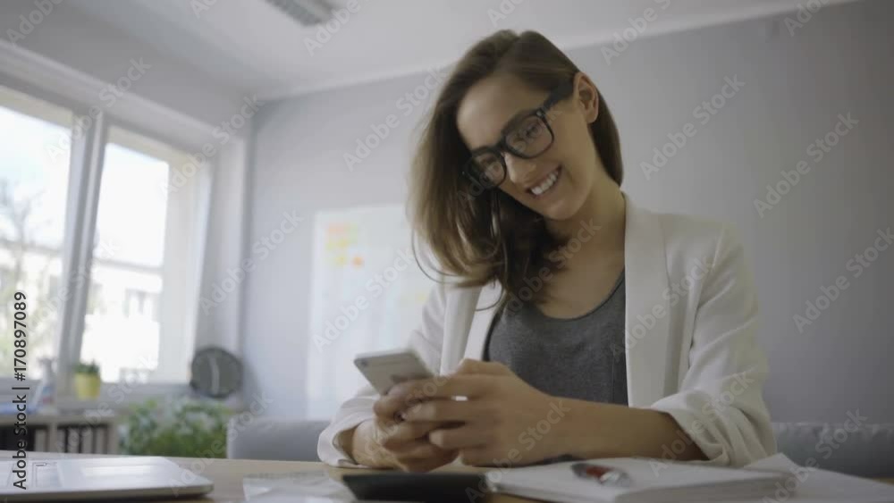 Beautiful young female entrepreneur sitting at desk in home office and using phone Beautiful young female entrepreneur sitting at desk in home office and using phone
