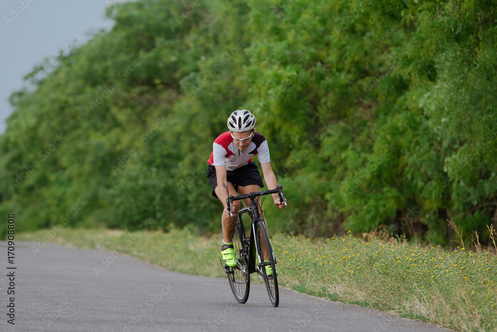Bicycle racer in helmet and sportswear training alone on empty country road, fields and trees background Bicycle racer in helmet and sportswear training alone on empty country road, fields and trees background