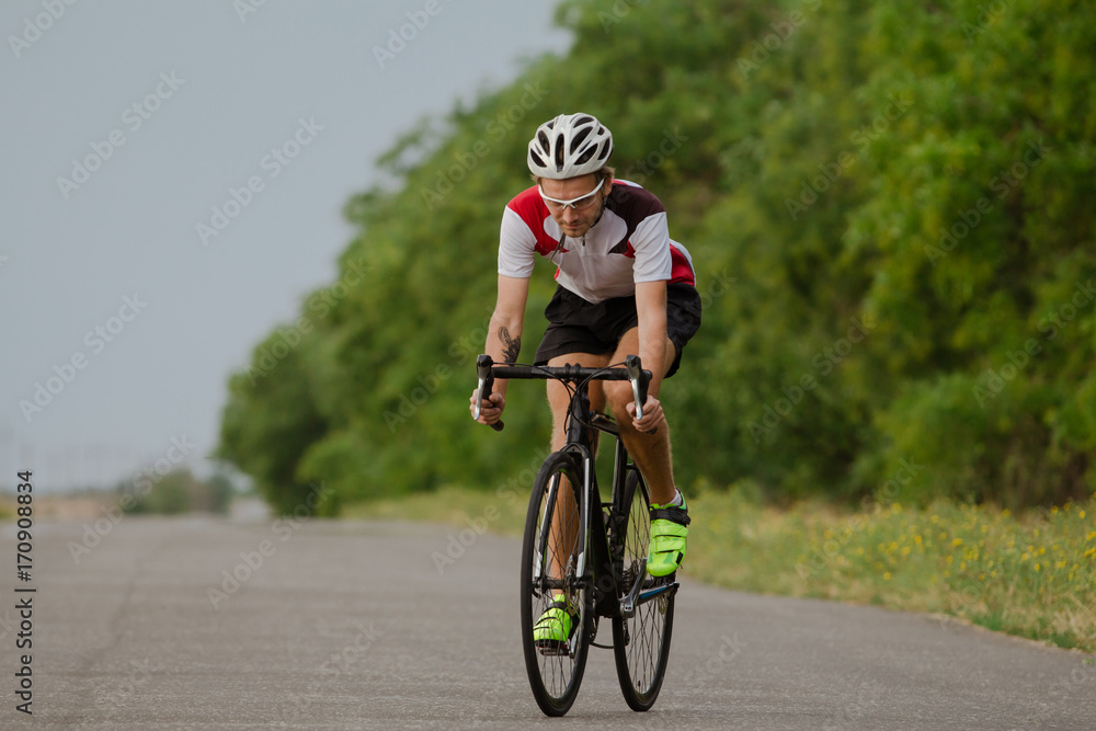 Fototapeta premium Bicycle racer in helmet and sportswear training alone on empty country road, fields and trees background 