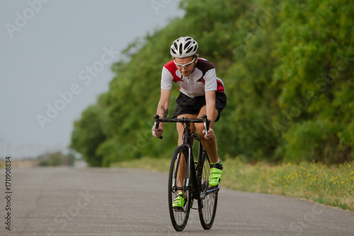 Bicycle racer in helmet and sportswear training alone on empty country road, fields and trees background 