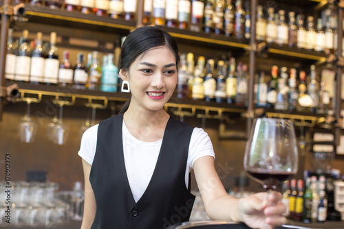 Young Asian Woman Waiter Restaurant Catering Service Happy Emotion. Woman Waiter Present Red-Wine for Customer at Bar. Woman with Wine in Bar Concept.