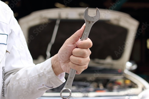 Close up hands of professional young mechanic man holding wrench with car in open hood at the garage background. Auto repair service.