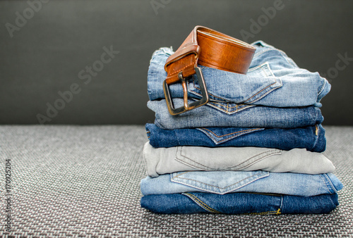 Stack of different jeans, rolled men's leather belt on gray background.