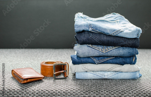 Stack of different jeans, rolled men's leather belt and wallet on gray background.