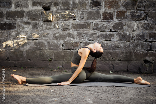Beautiful young woman practicing yoga hanumanasana by a brick wall. Yoga concept.